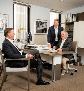 Three men in business attire have a discussion in a modern office; one is seated at a desk, another sits across from him, and the third stands between them, smiling.