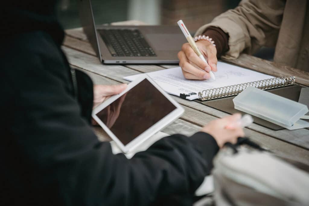 Two people work at a table with a tablet, laptop, and papers. One person writes in a notebook while the other holds a tablet.