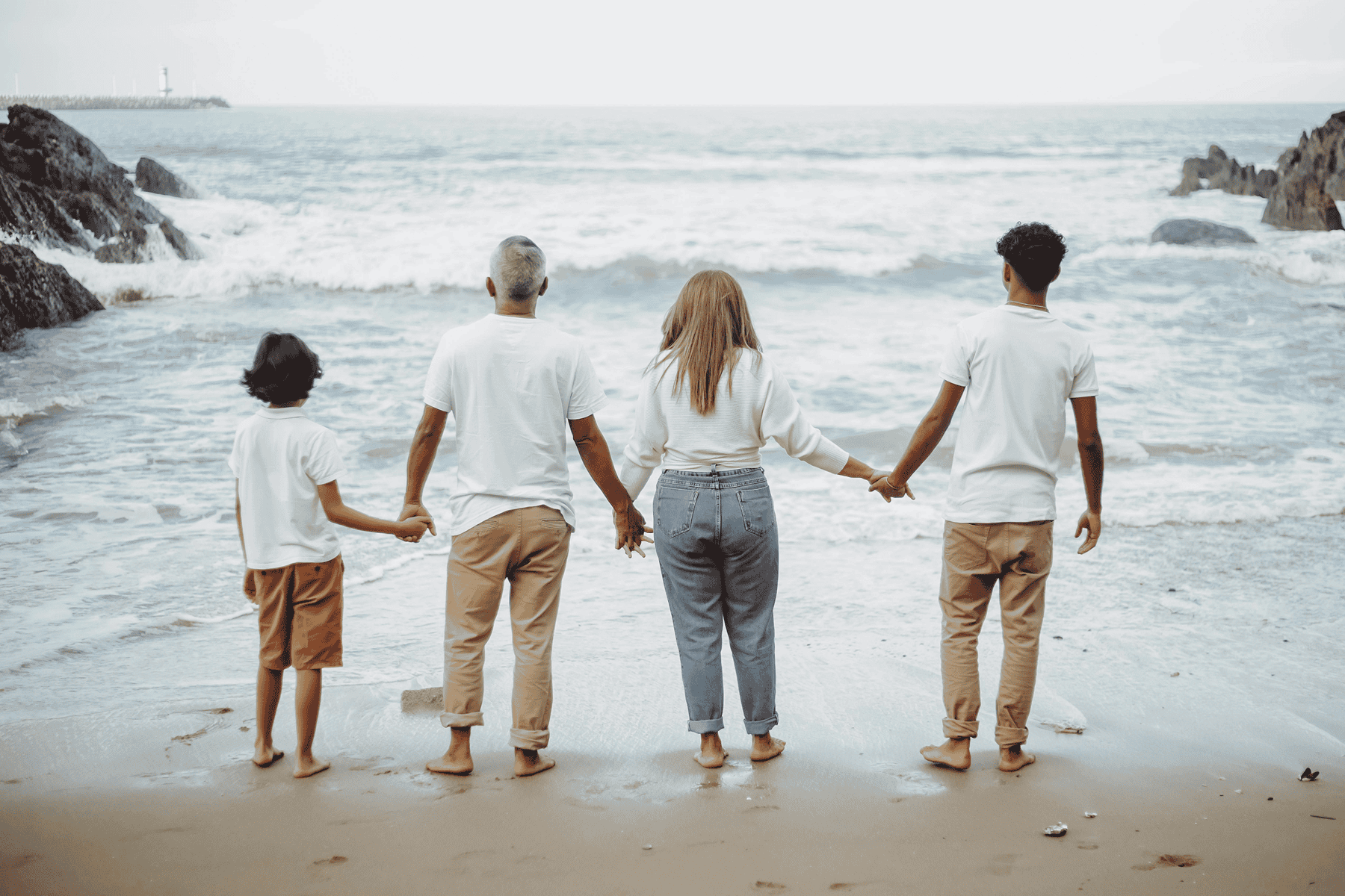 Four people stand barefoot on the beach, holding hands and facing the ocean. Two adults are in the center with two children on either side.