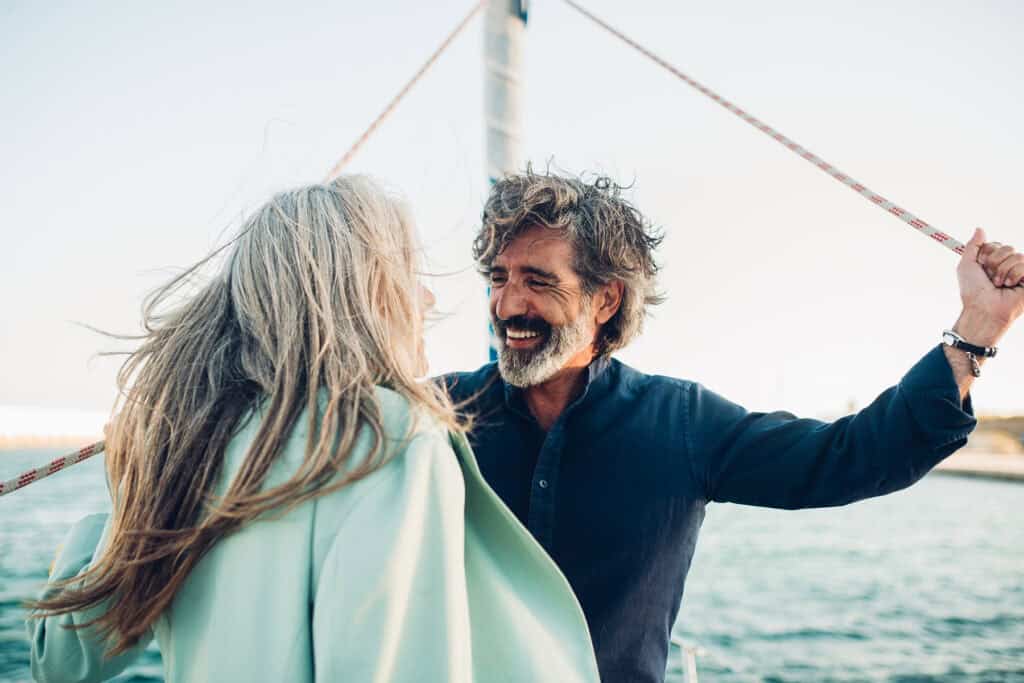 An older man and woman stand on a boat, smiling and facing each other, with water and sky in the background.