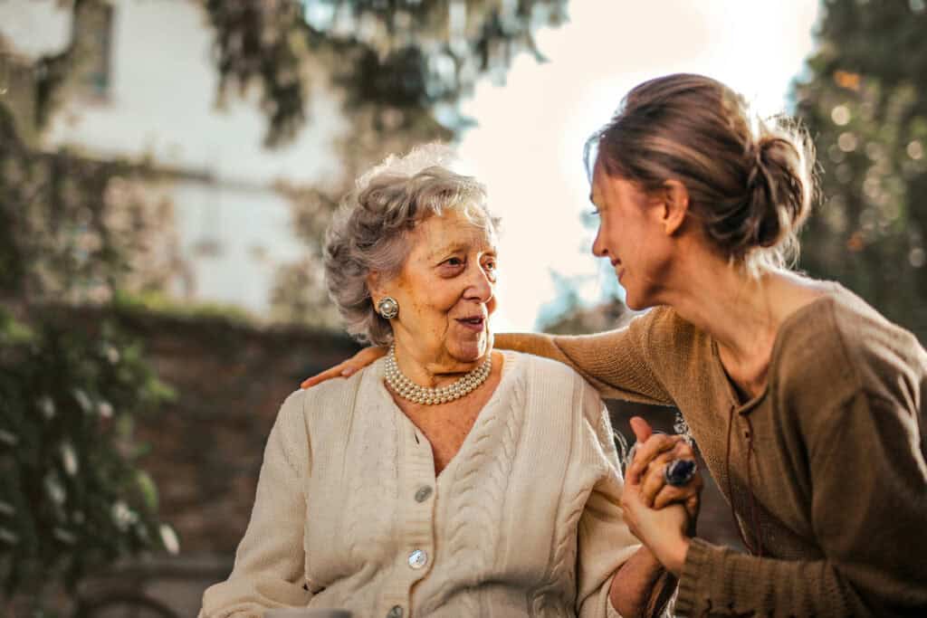 An elderly woman and a younger woman sit outdoors, smiling and holding hands, with greenery and sunlight in the background.
