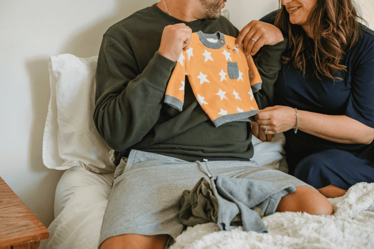 A man and woman sit on a bed smiling while holding up a small, orange, star-patterned baby sweater.