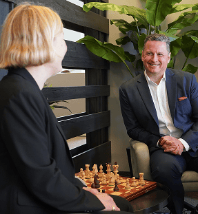 Two people in business attire sit across from each other, smiling, with a chessboard on the table between them and large green plants in the background.