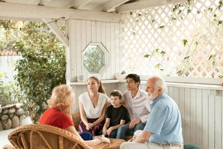 Five people, including two older adults, a man, a woman, and a child, are sitting together on wicker furniture in a bright, covered outdoor patio area.