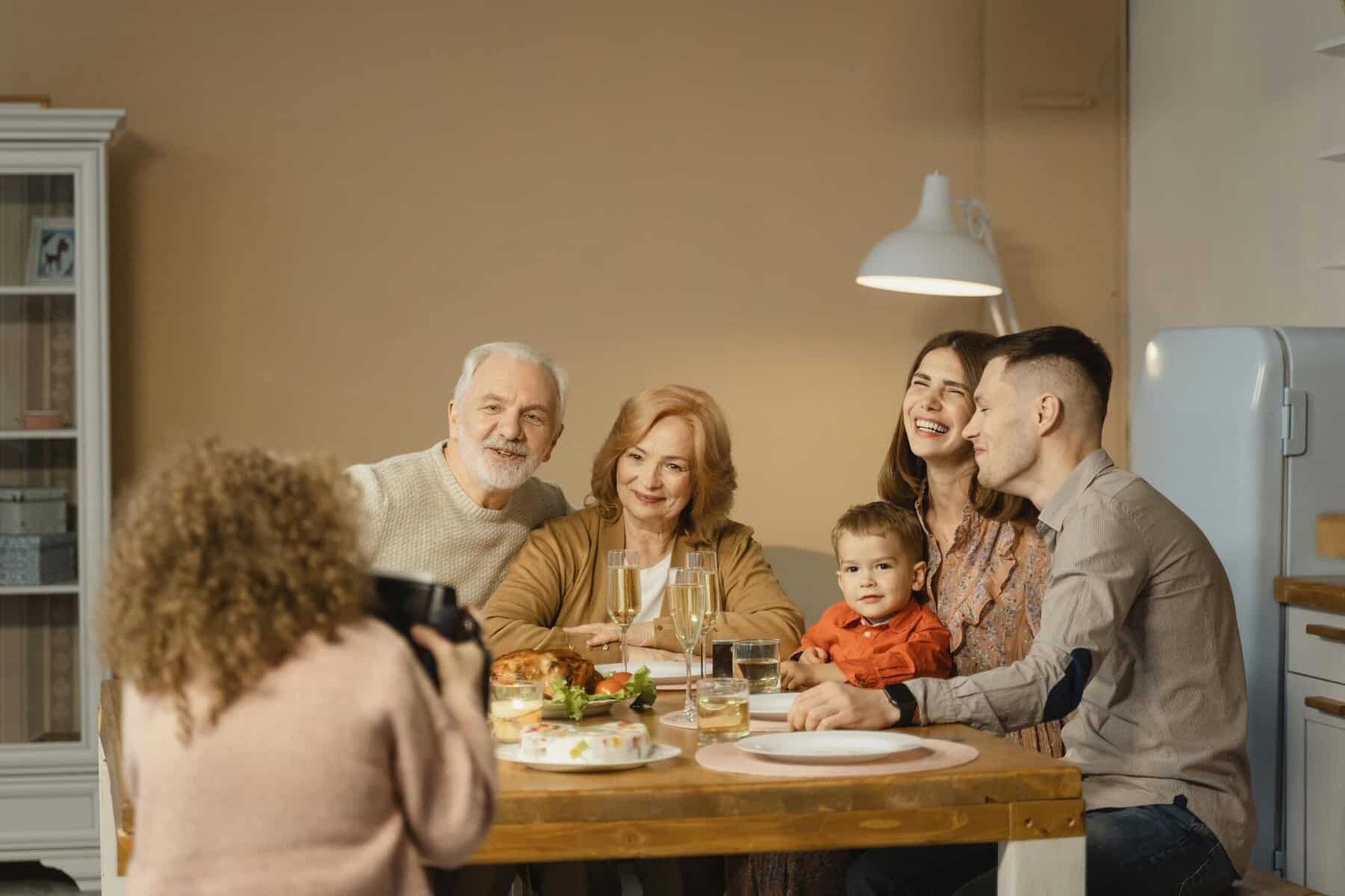 A group of five people, including a child, sit around a dining table with food and drinks, posing for a photo taken by a person with curly hair.