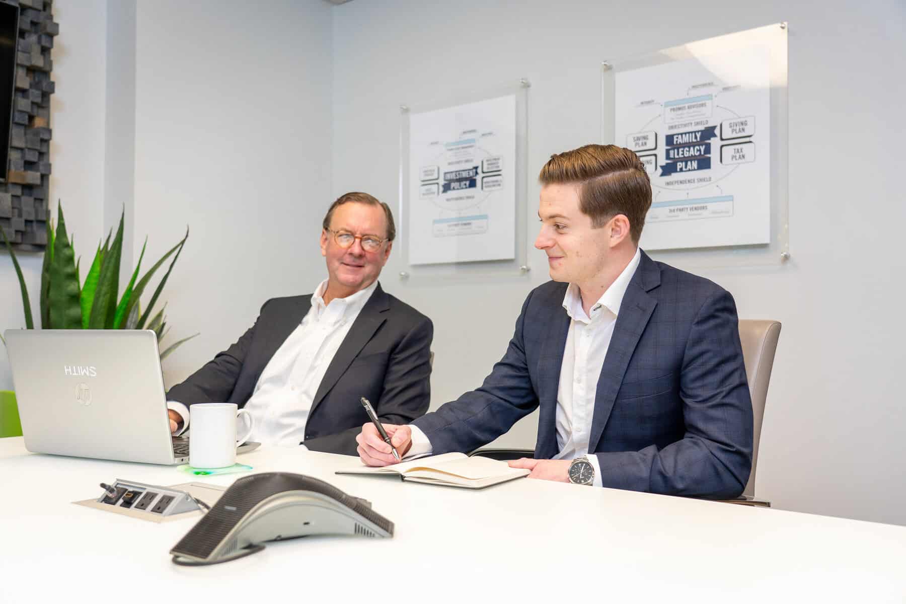 Two men in business suits sit at a conference table; one is using a laptop while the other writes in a notebook. A speakerphone and documents are on the table.